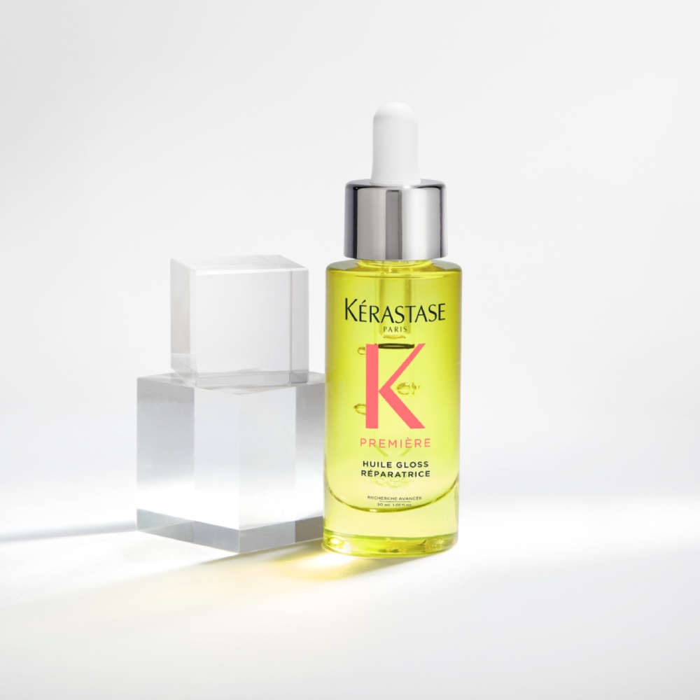 Première Huile gloss réparatrice hair oil is displayed on a pure grey background with transparent cubes. The oil repairs shine and split ends while neutralizing dullness.